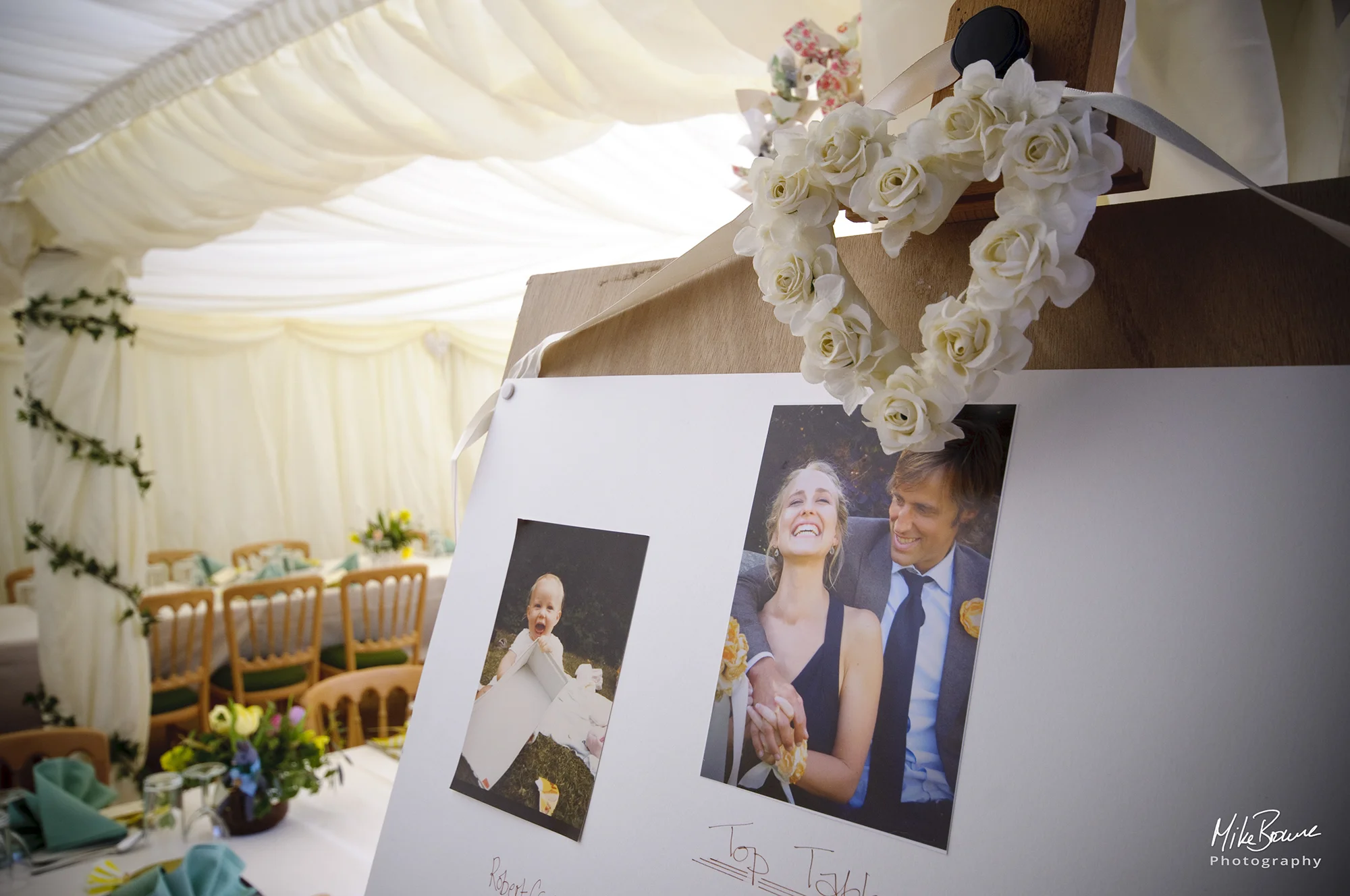 Photo board in a marquee with heart shaped bouquet of white roses and pictures of a young man and woman displayed on it