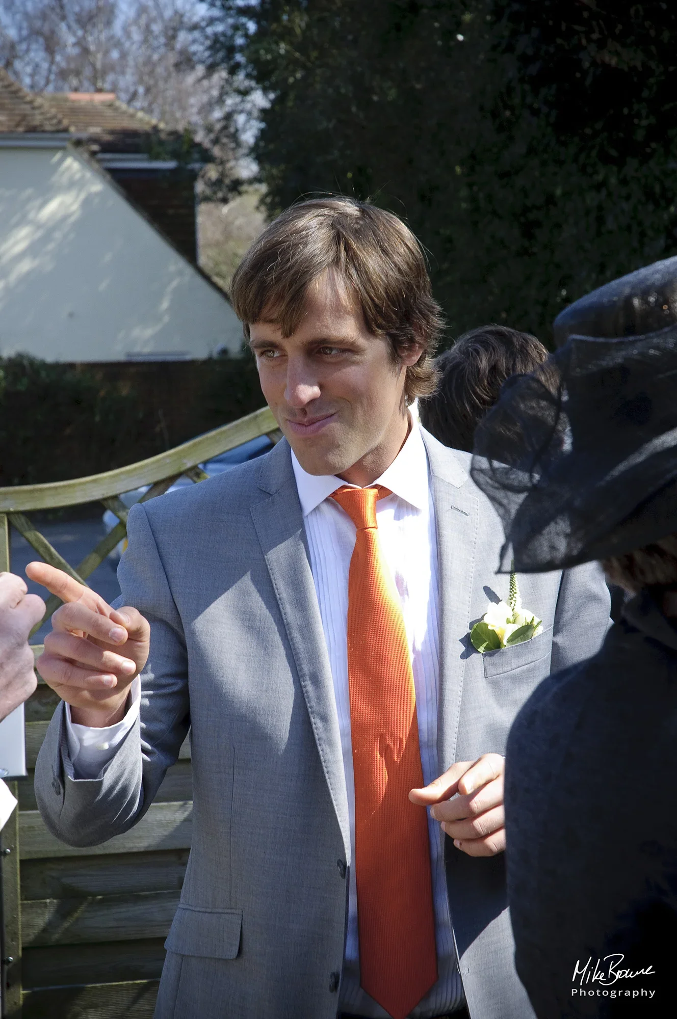 Man wearing orange tie and grey jacket with yellow rose button hole pointing at someone and smiling