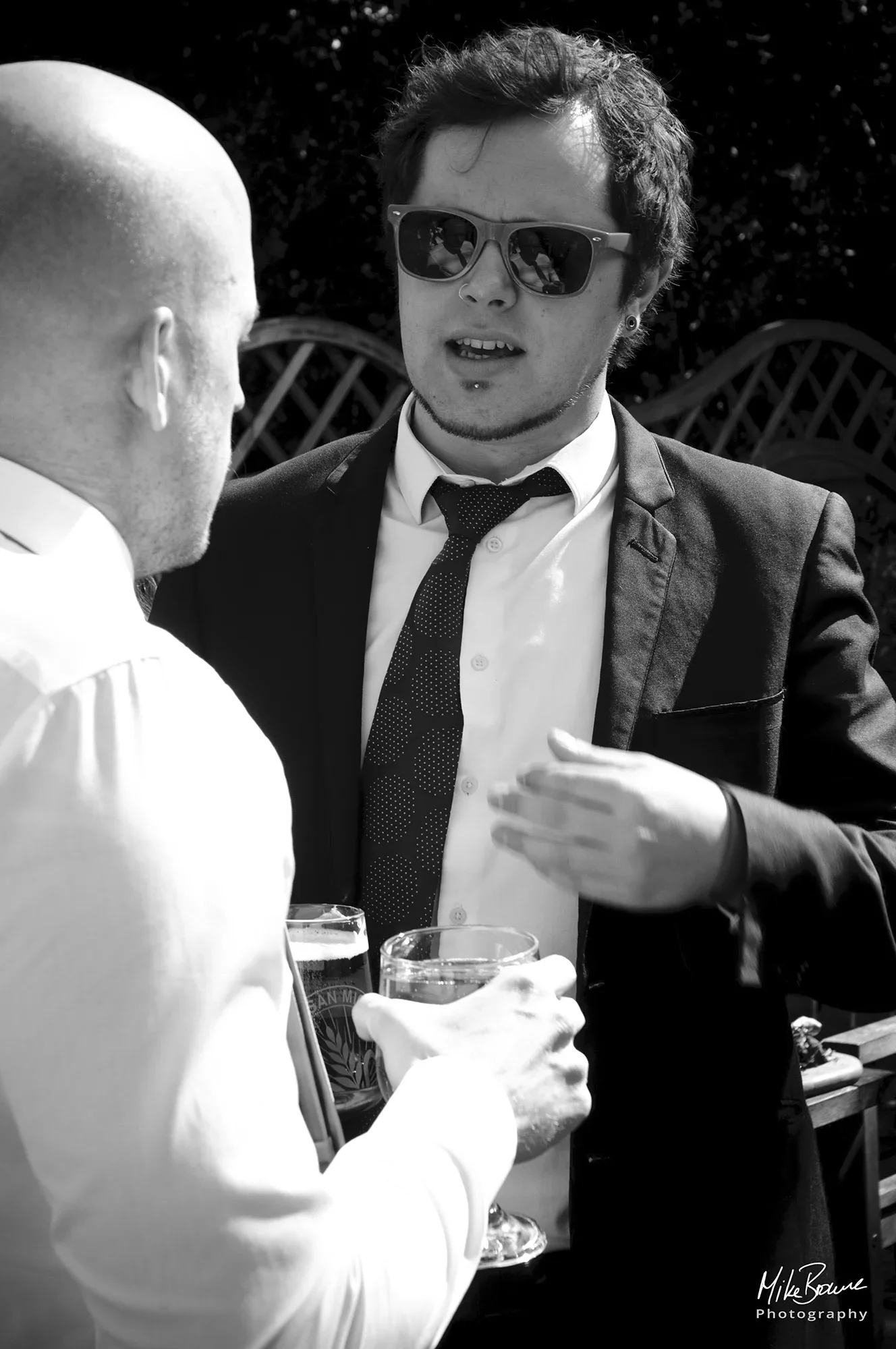 Man in sunglasses and dark suit talking to a man in a white shirt holding beers