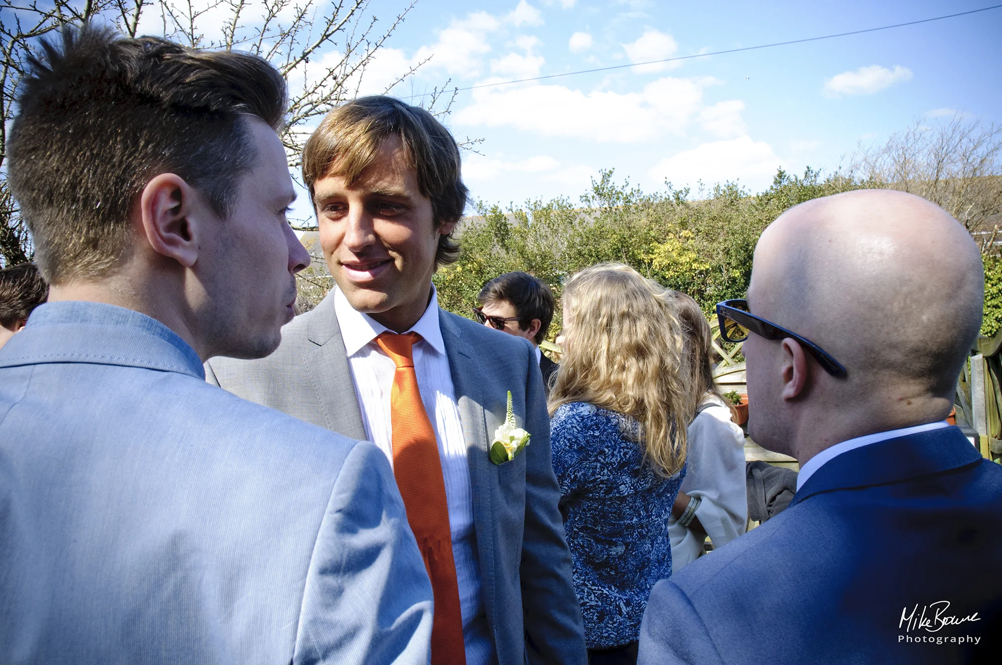 Man in grey jacket and orange tie talking to two friends in a garden