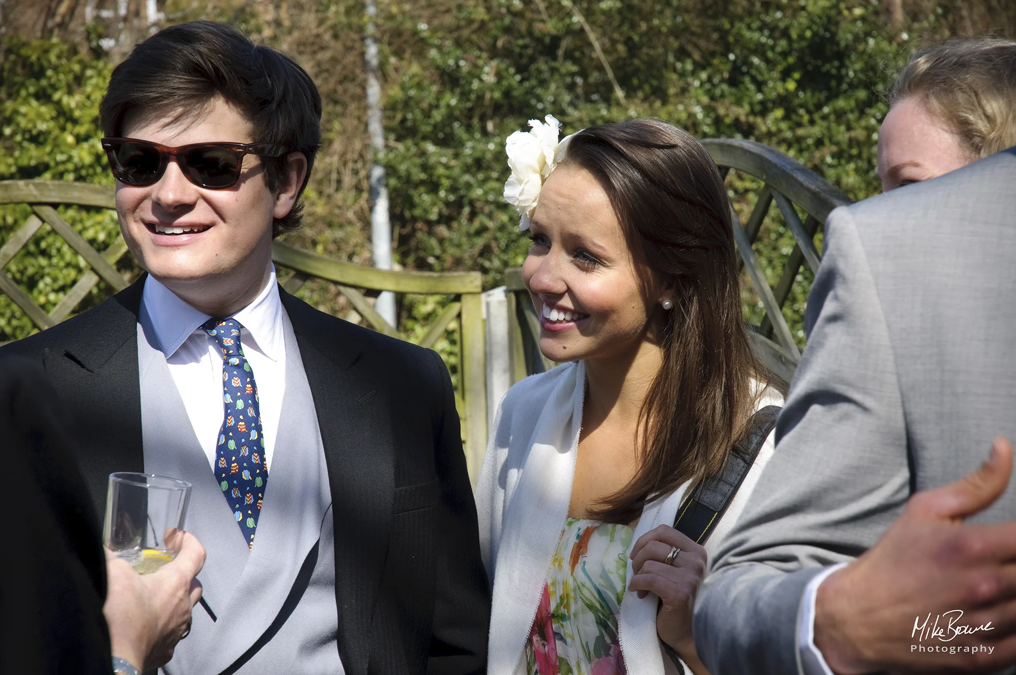 Man in morning suit and a woman with white flower in her hair talking to woman holding a gin and tonic in a pub garden