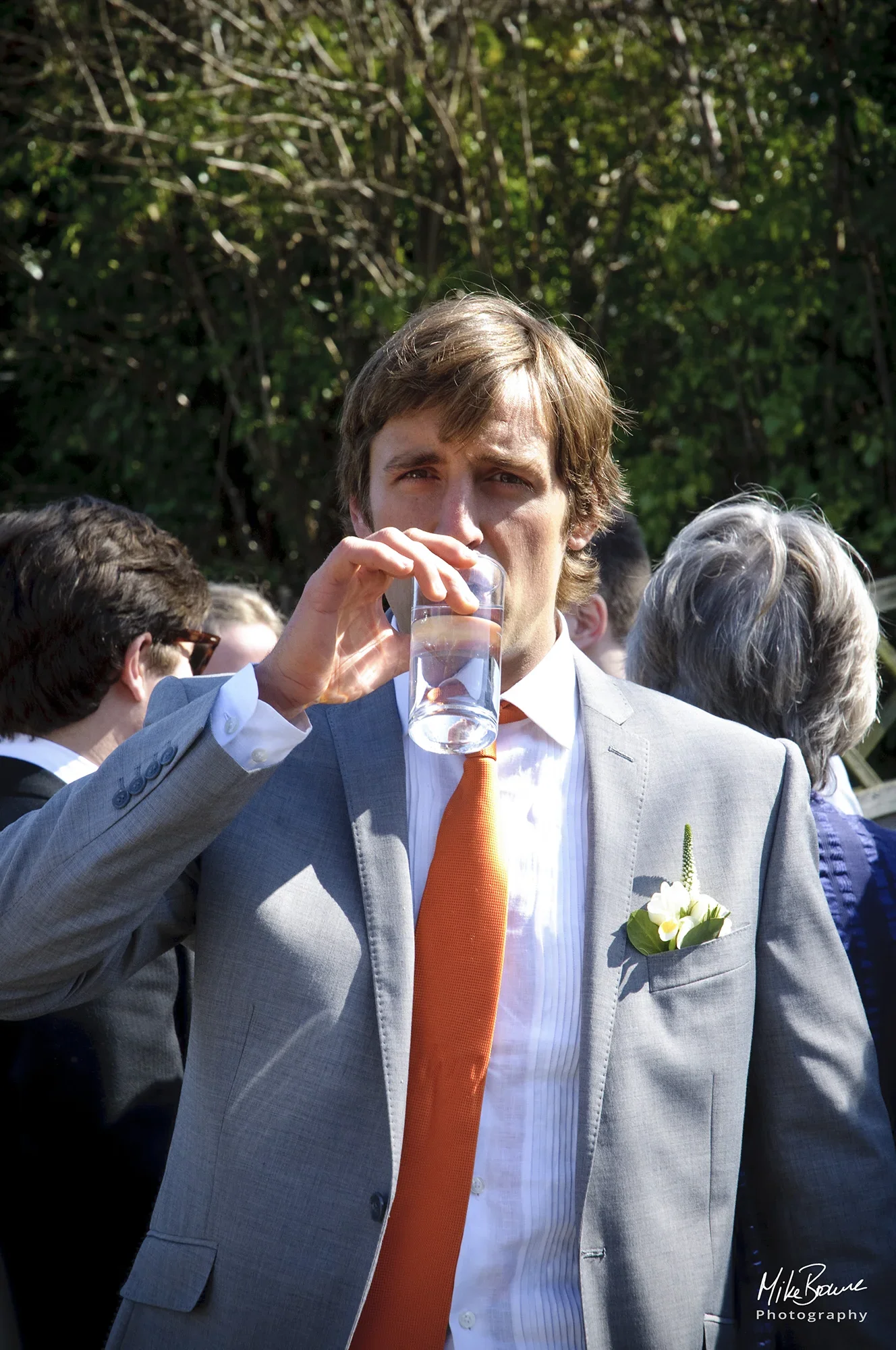 Bridegroom wearing grey suit, orange tie and yellow rose button hole raising a glass to his lips