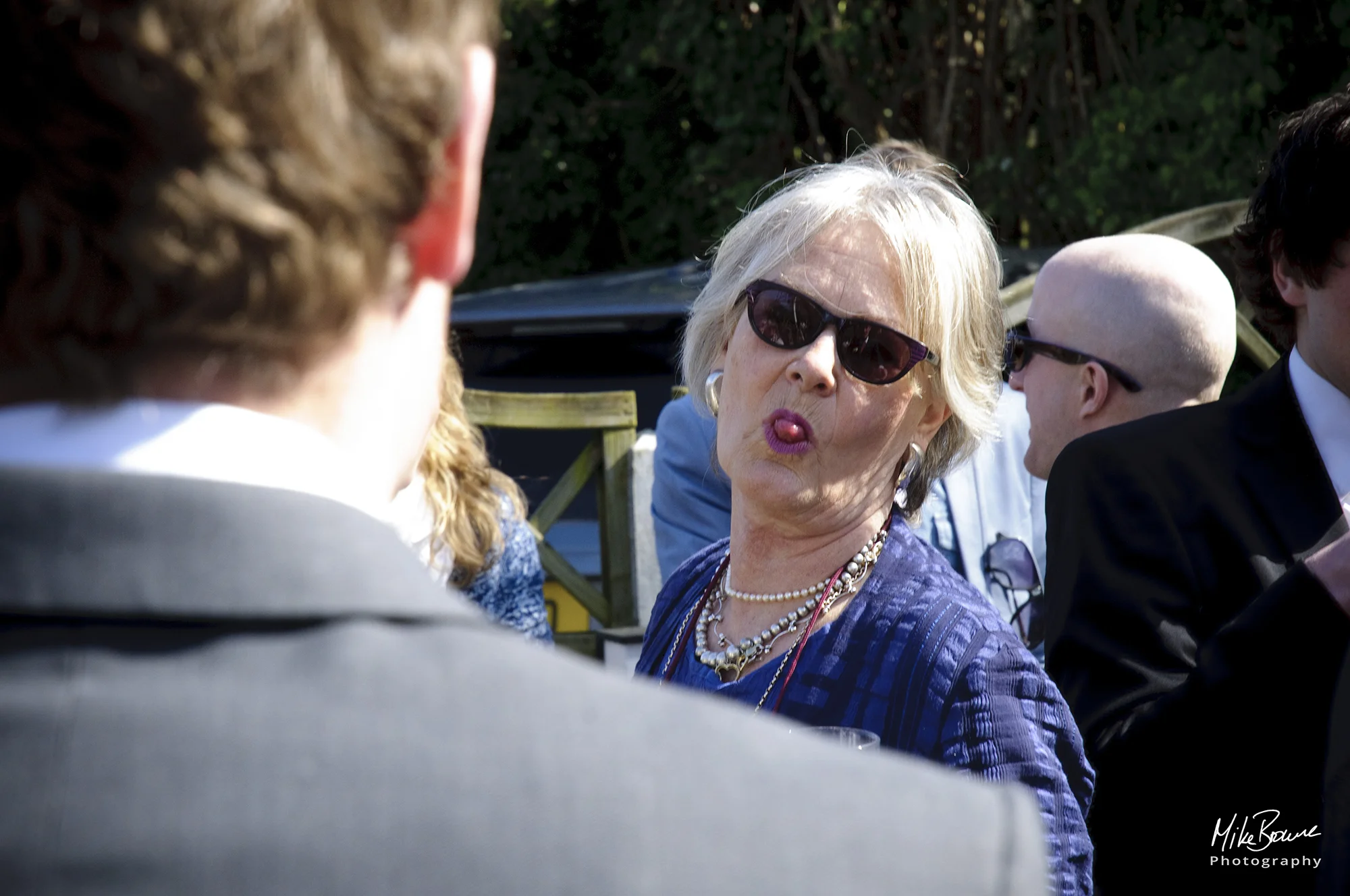 Older lady wearing sunglasses and necklaces poking her tongue out at the camera