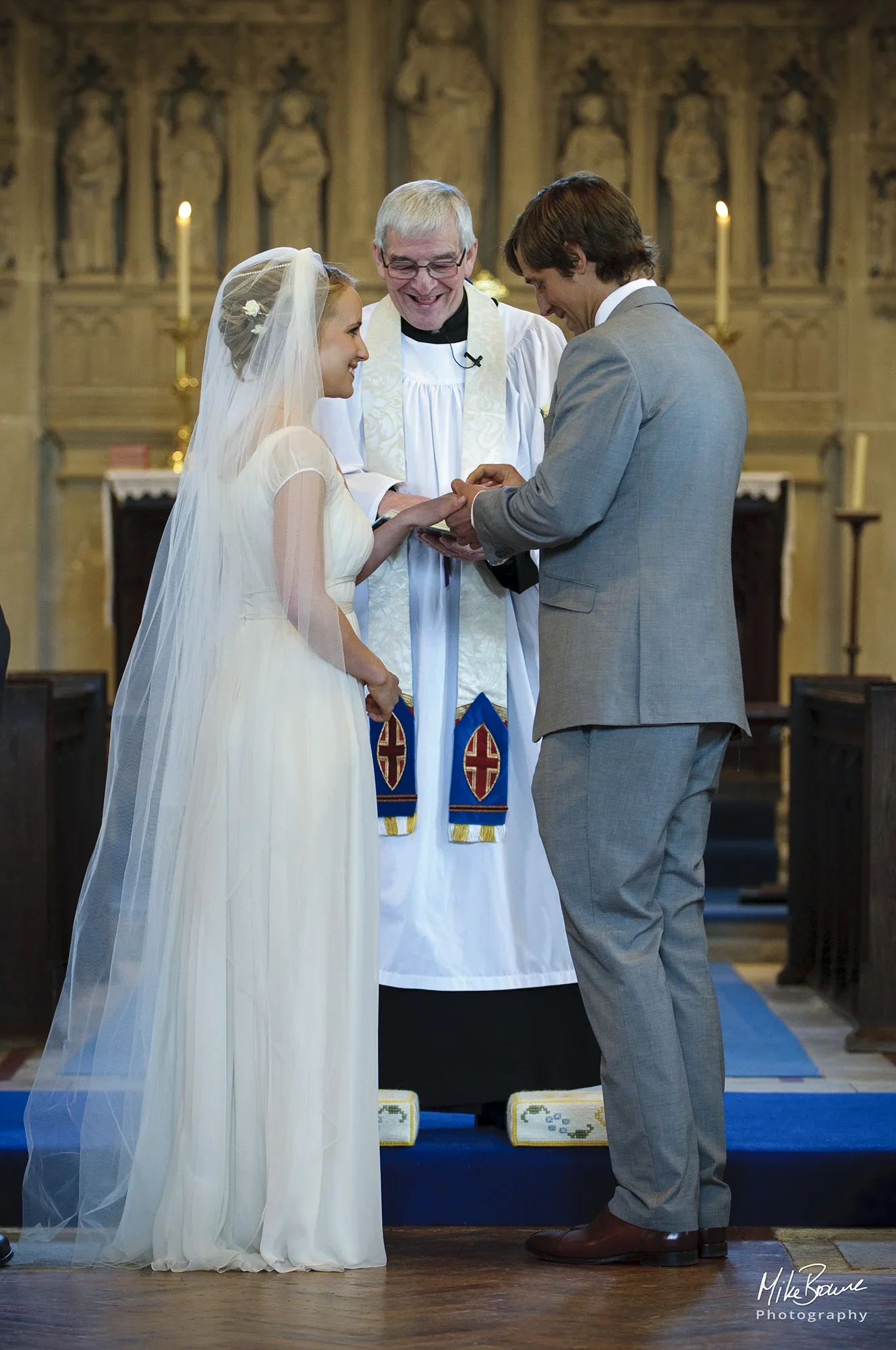 Bridegroom placing a wedding ring on his brides finger in front of a smiling vicar